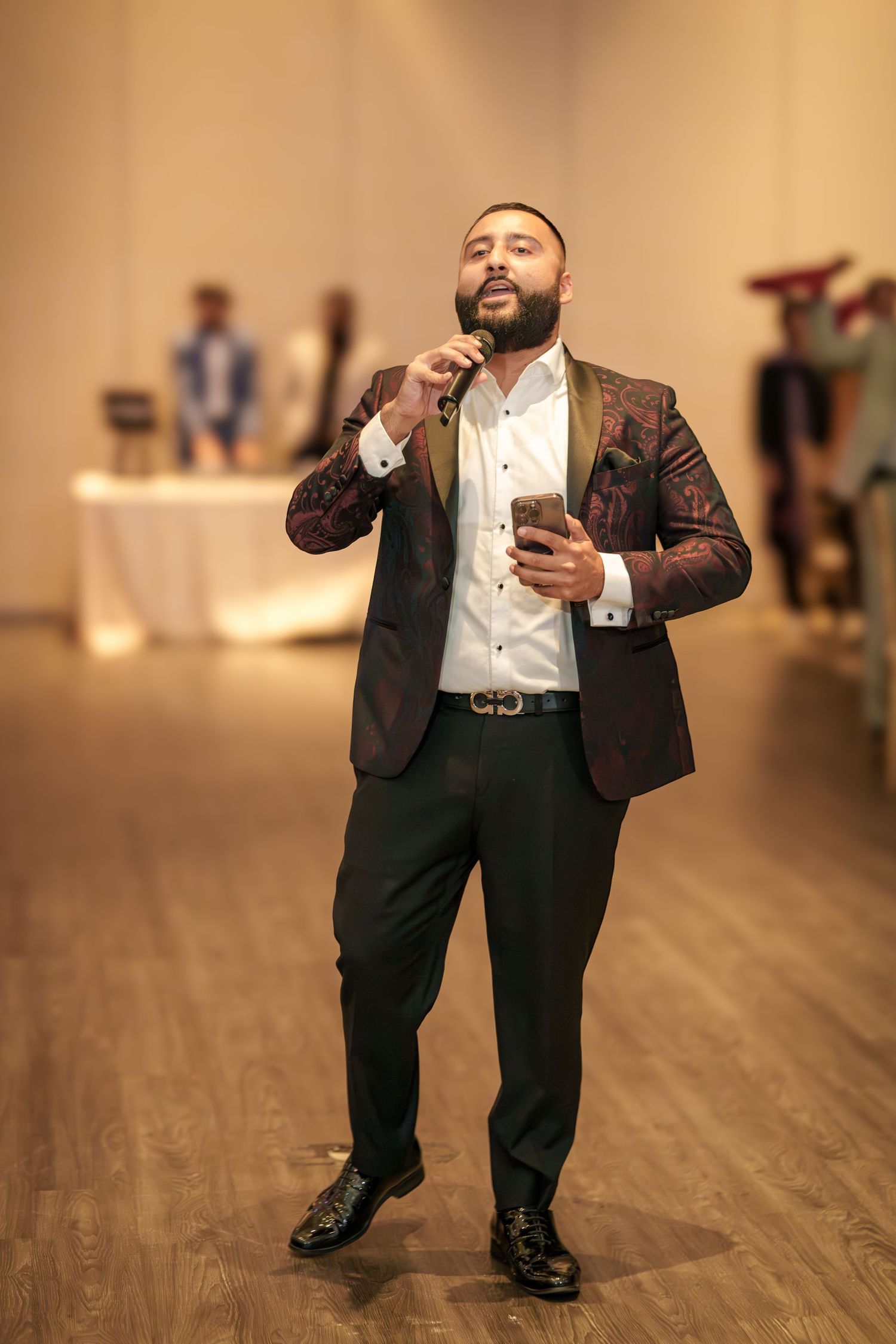 Reception guest in burgundy blazer holds microphone while speaking at wedding event.