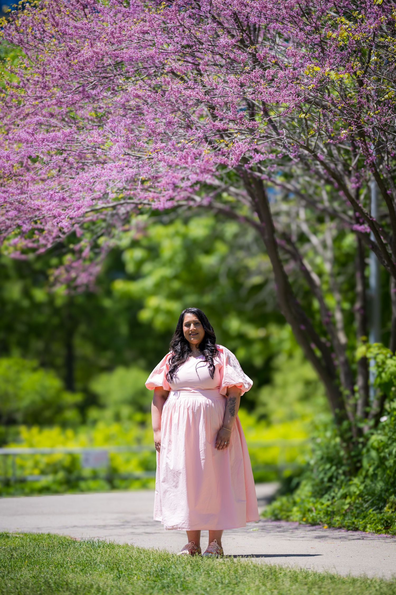 Smiling pregnant woman in pink midi dress poses near vibrant pink flowering redbud tree.