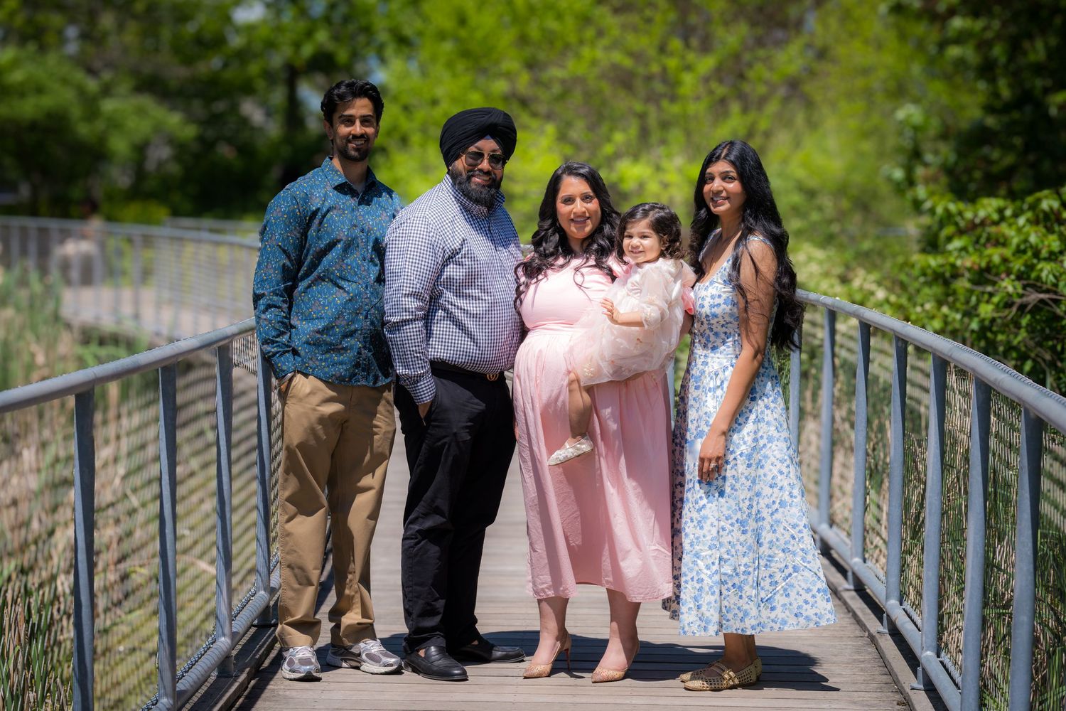 Family of five posing on wooden bridge in sunny park, including pregnant woman in pink dress.