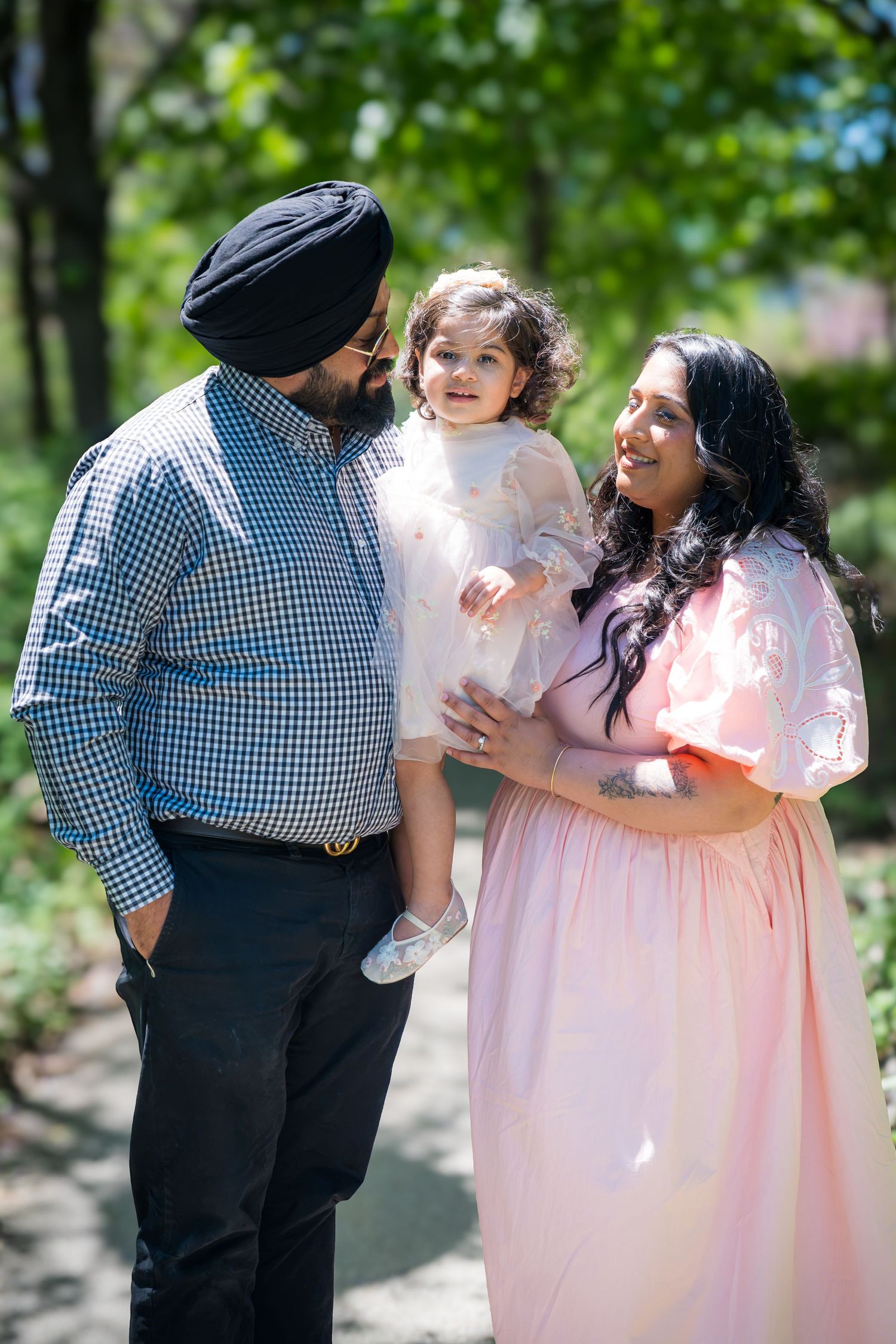 A Sikh couple smiles at their toddler daughter in a white dress in a sunny green park.