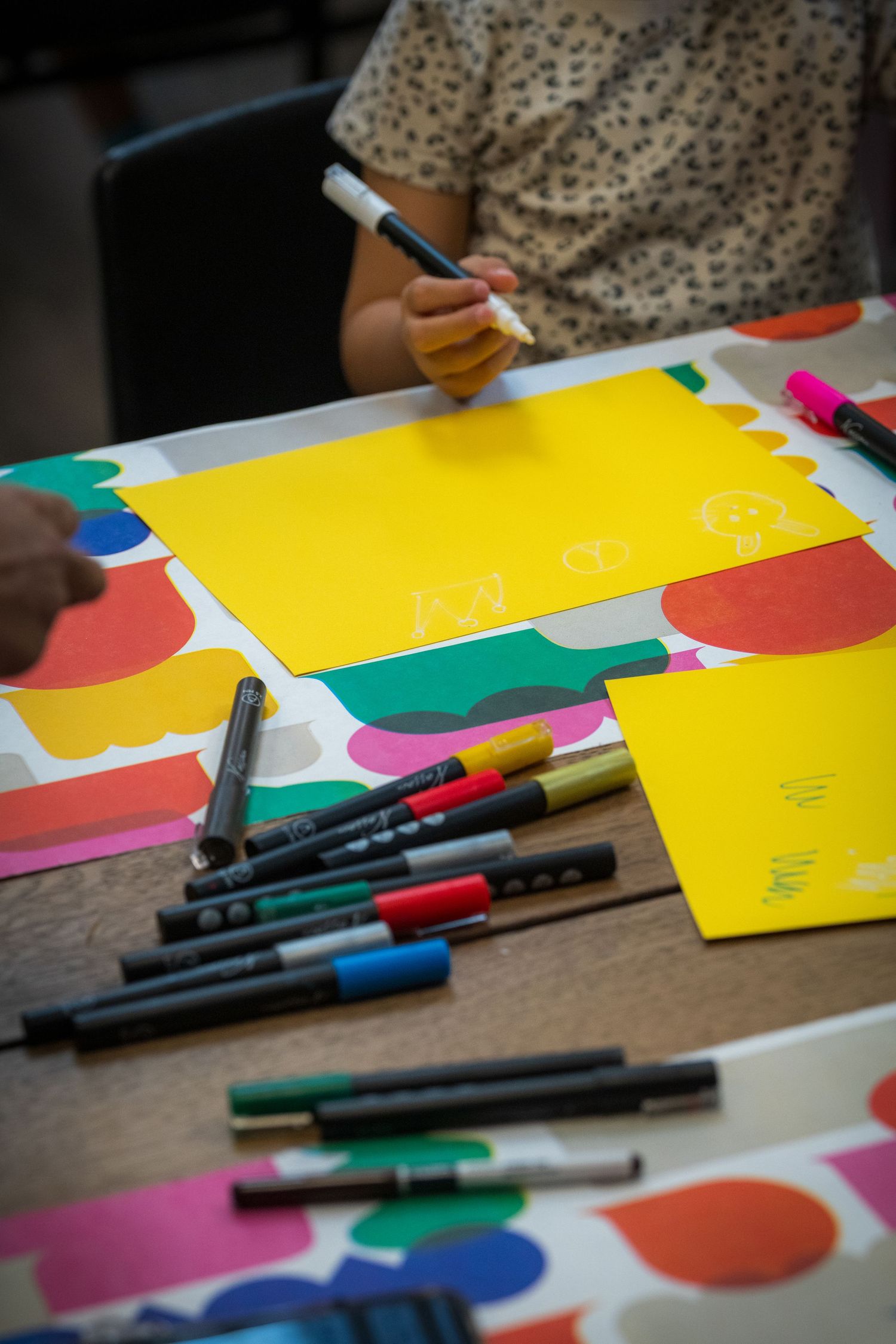Young child writing on yellow paper with markers scattered on a colorful table mat.