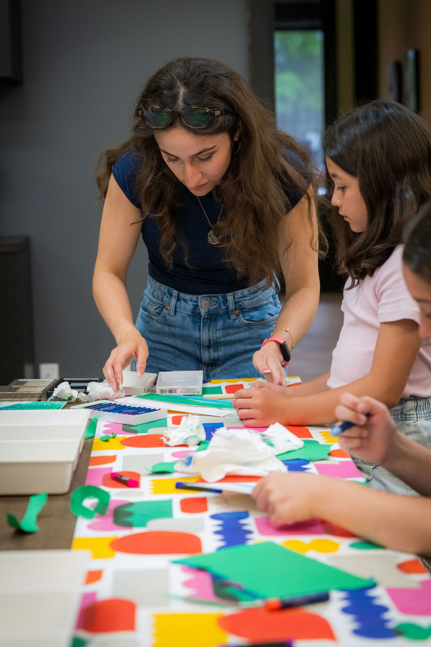 Female teacher leans over table helping children with colorful paper collage art project.