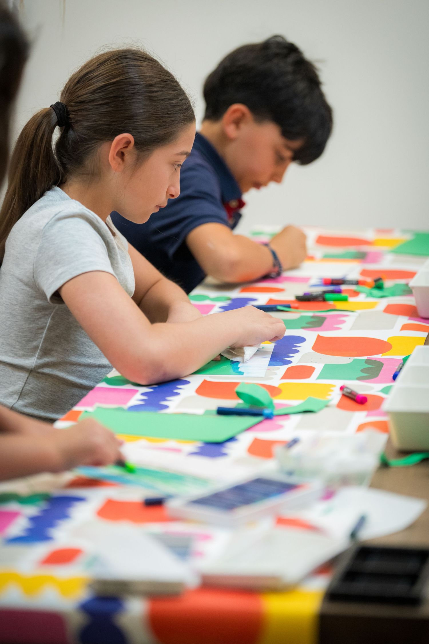Children engaged in a colorful art project at a table covered with bright paper cutouts and markers in a classroom.