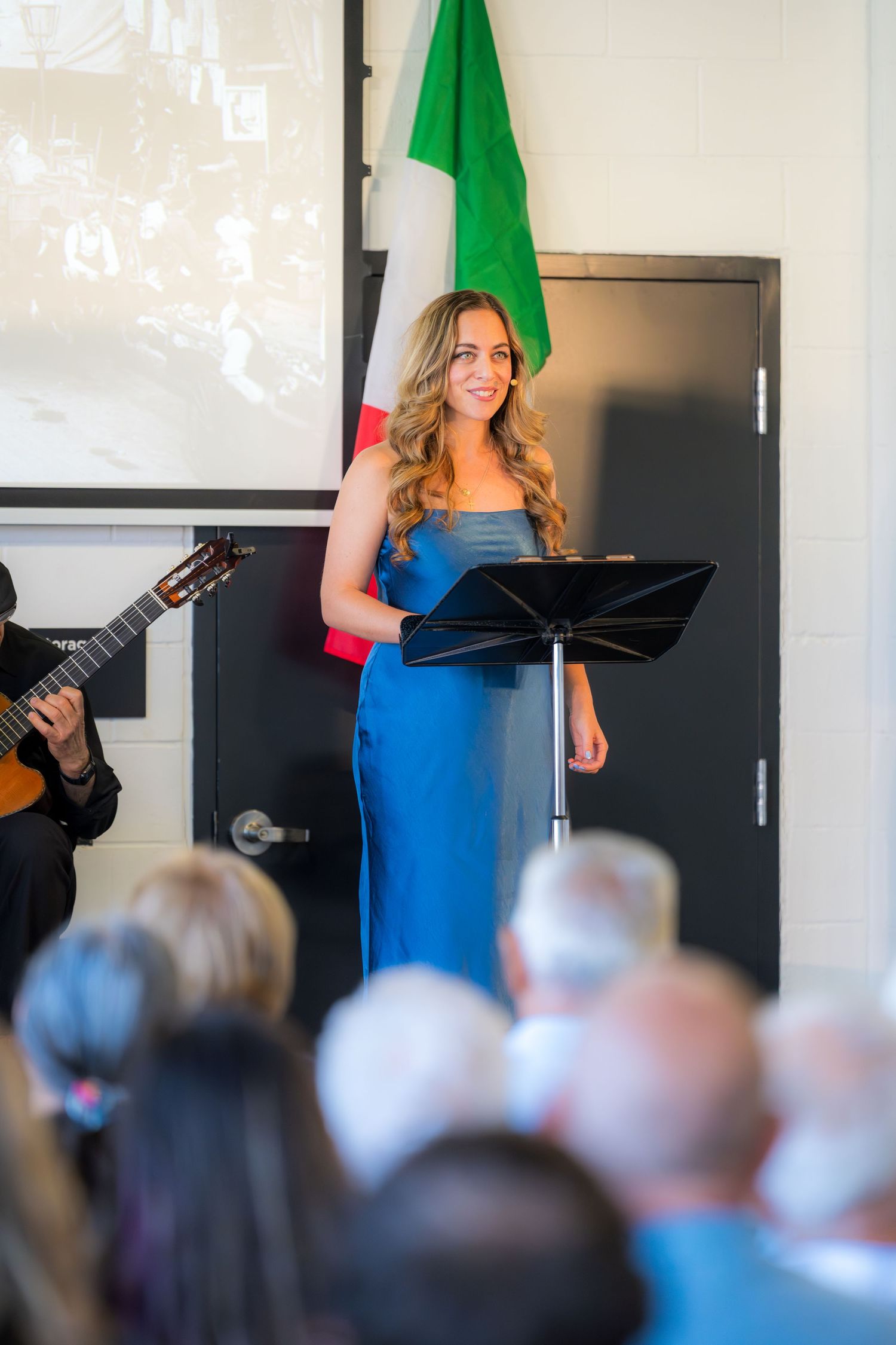 Young woman in blue dress presenting at music stand with Italian flag and guitarist nearby.