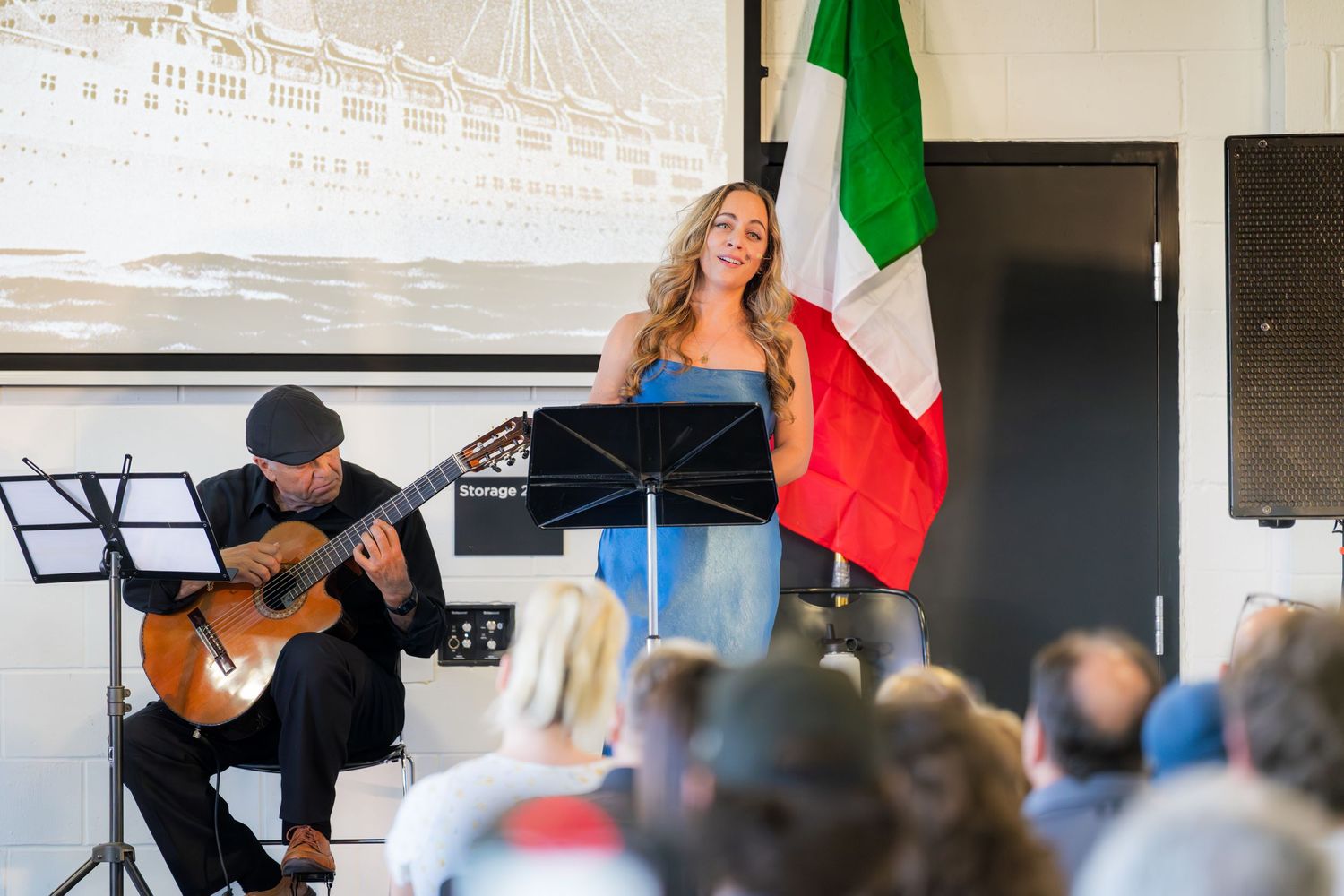 Female vocalist performing with acoustic guitarist at Italian heritage event, Italian flag prominently displayed.