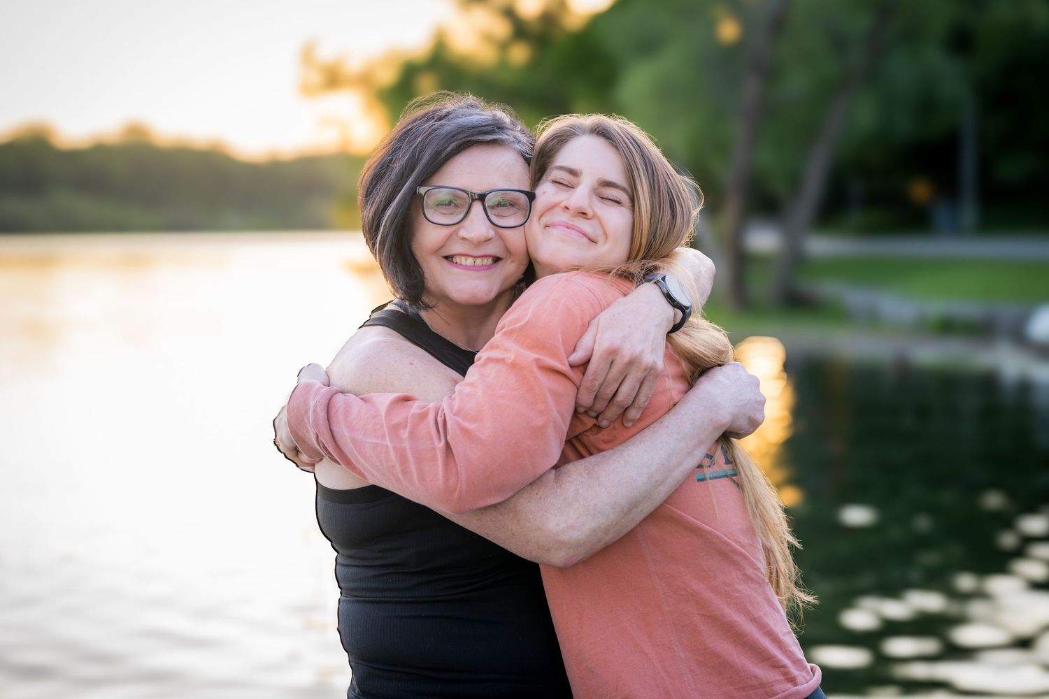 Two women sharing a warm joyful hug by a glowing sunset lakeside.