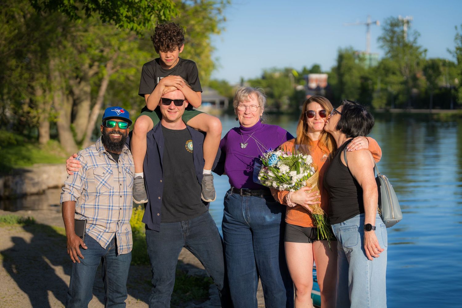 Group of six smiling people standing together near a riverbank on a bright sunny day.