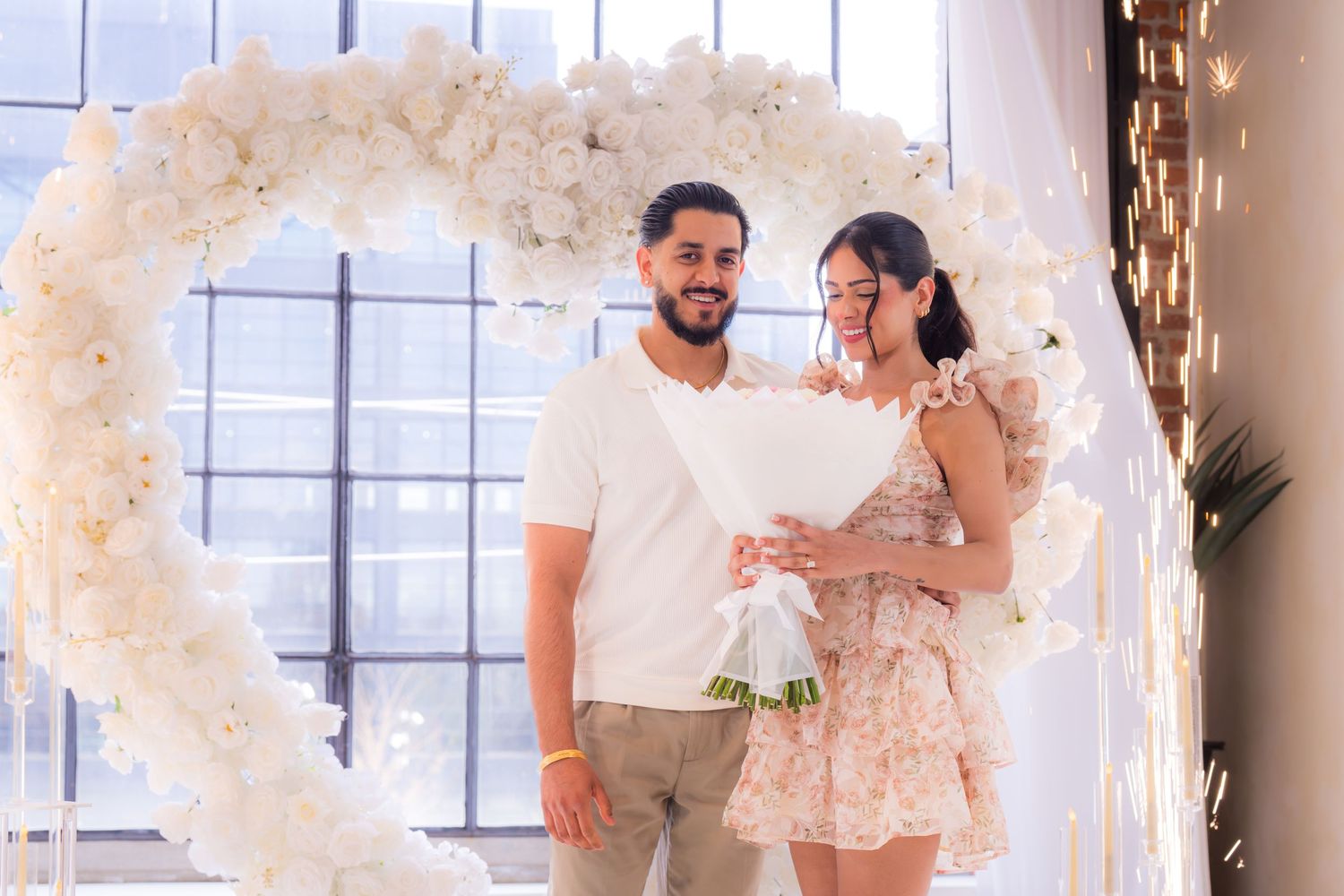 Couple reading letter together under white floral heart arch during engagement celebration.