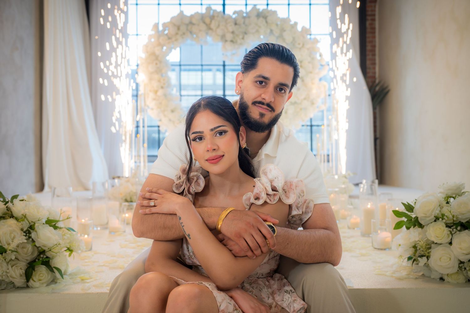 Newly engaged couple sits together in front of a glowing white floral heart arch with candles.