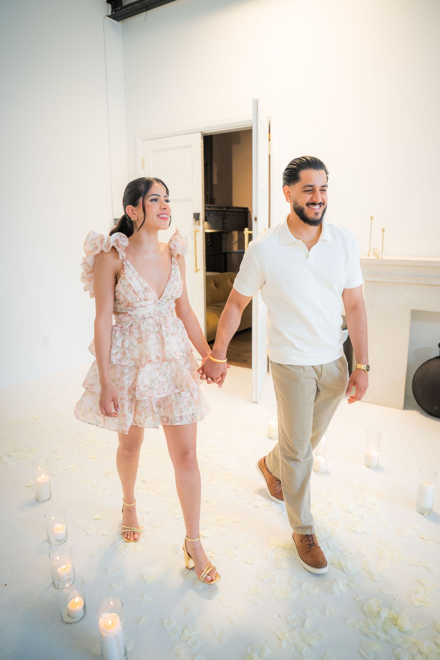 A joyful couple holds hands and walks through a romantic candlelit white room during a proposal setup.