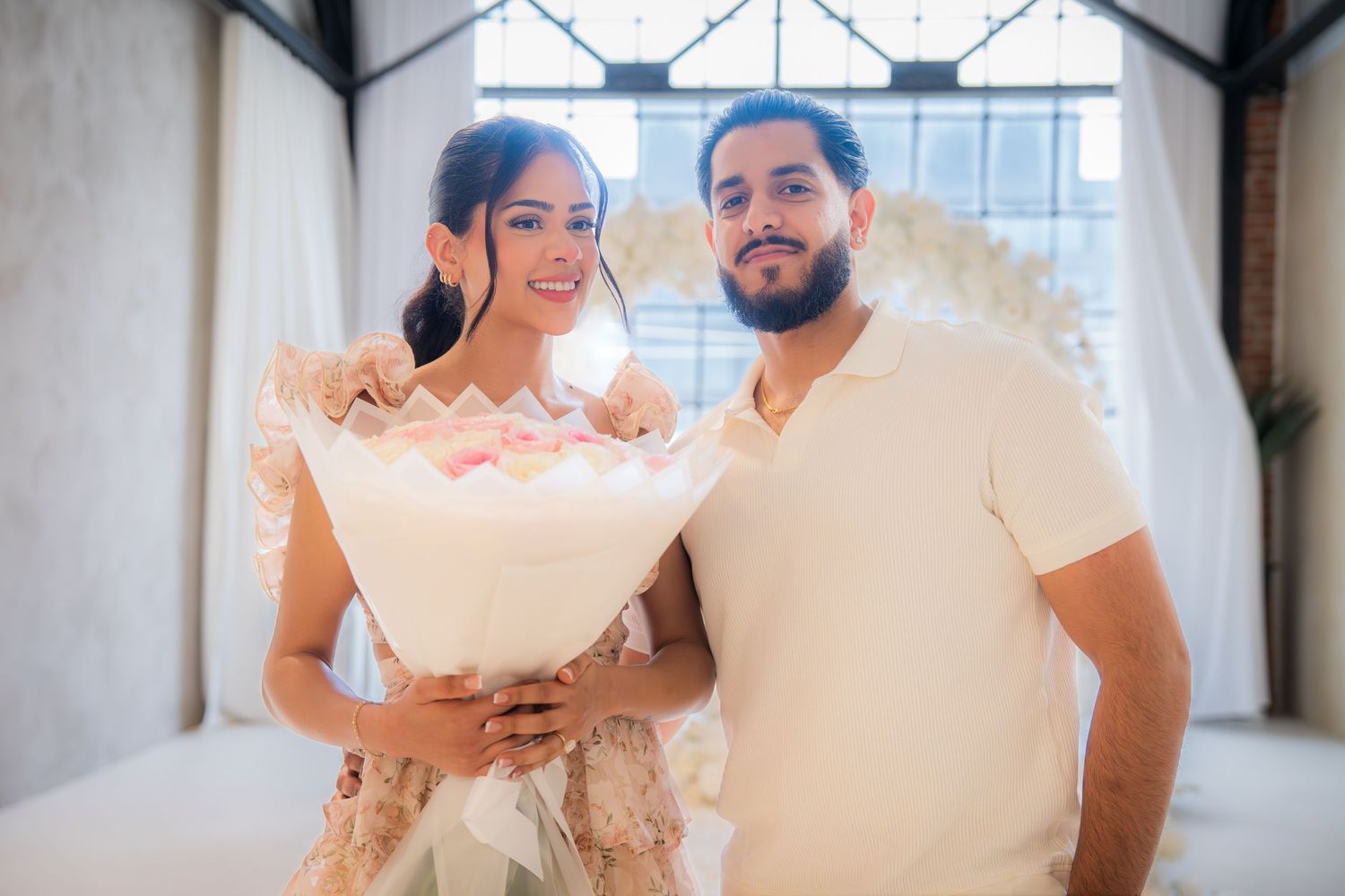 Couple smiling together holding a large floral bouquet in a bright white decorated room.
