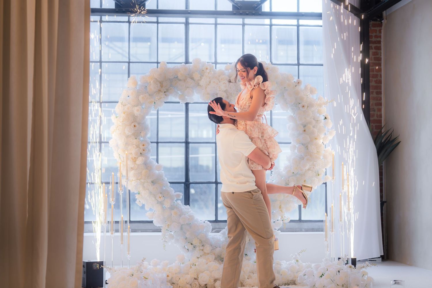 Man joyfully lifting his fiance in front of a white floral heart arch after a romantic marriage proposal.