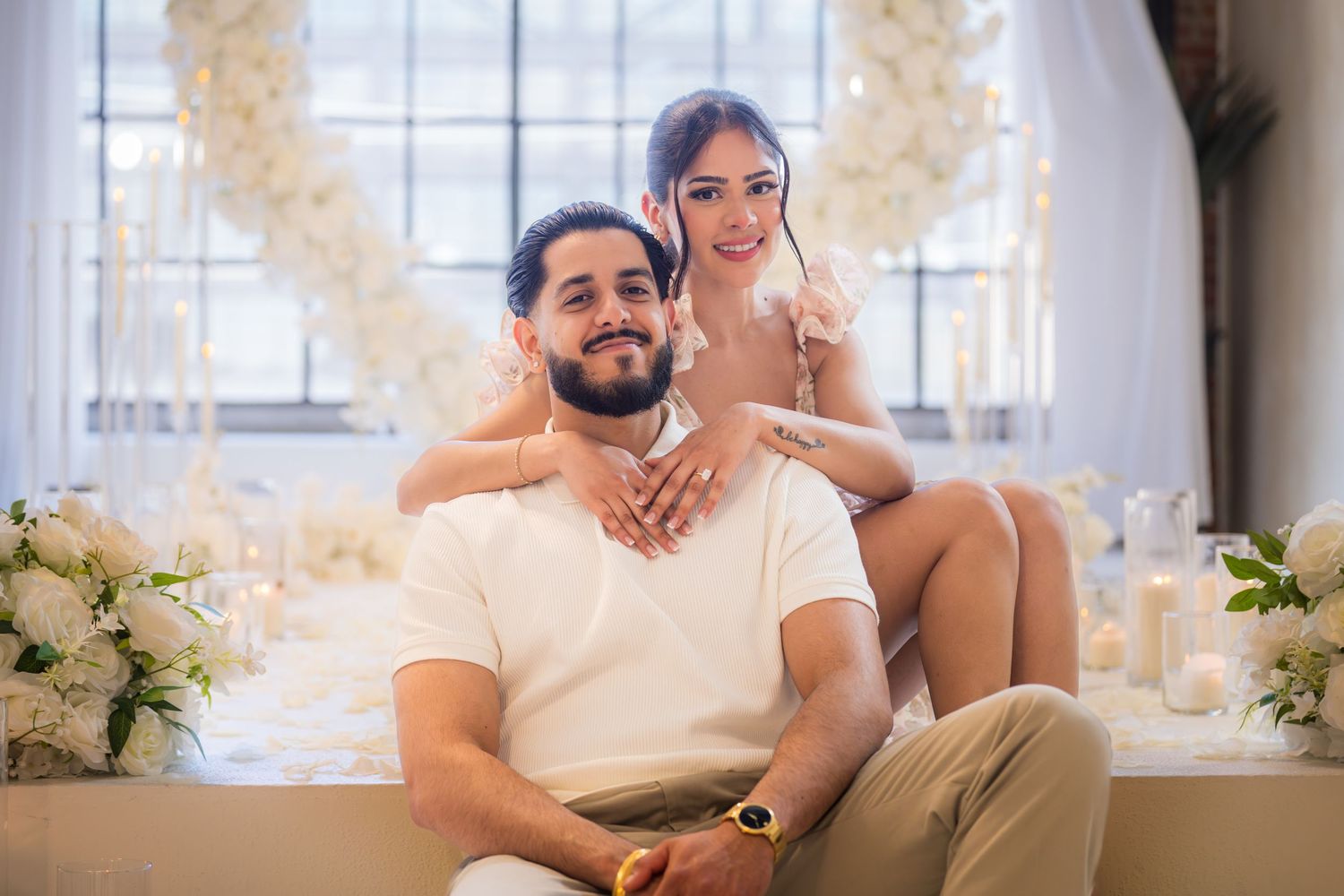 Engaged couple sitting together in front of white floral heart arch with candles and flower arrangements.