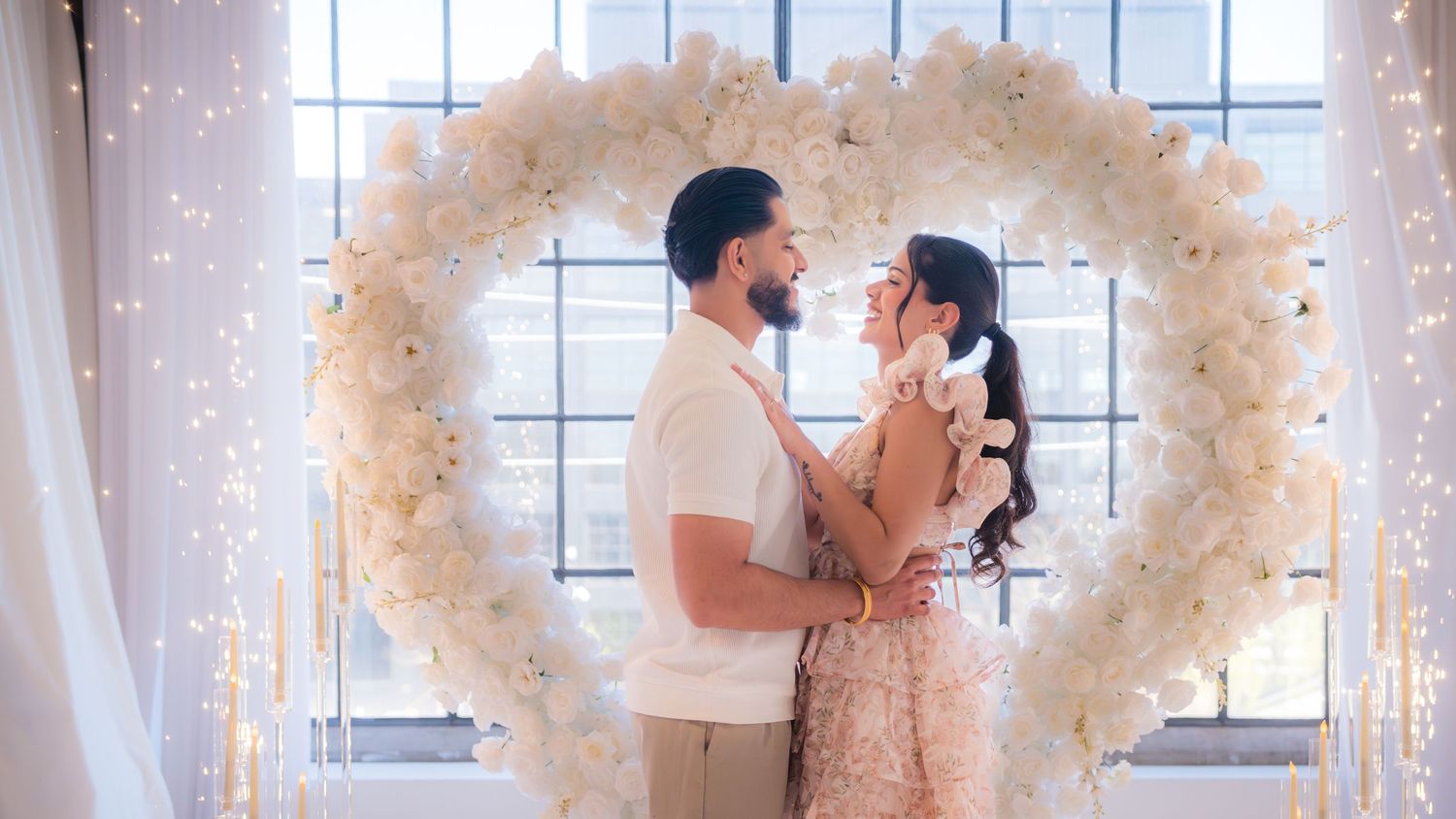 Couple sharing a romantic kiss under a large white floral heart arch with soft lighting.
