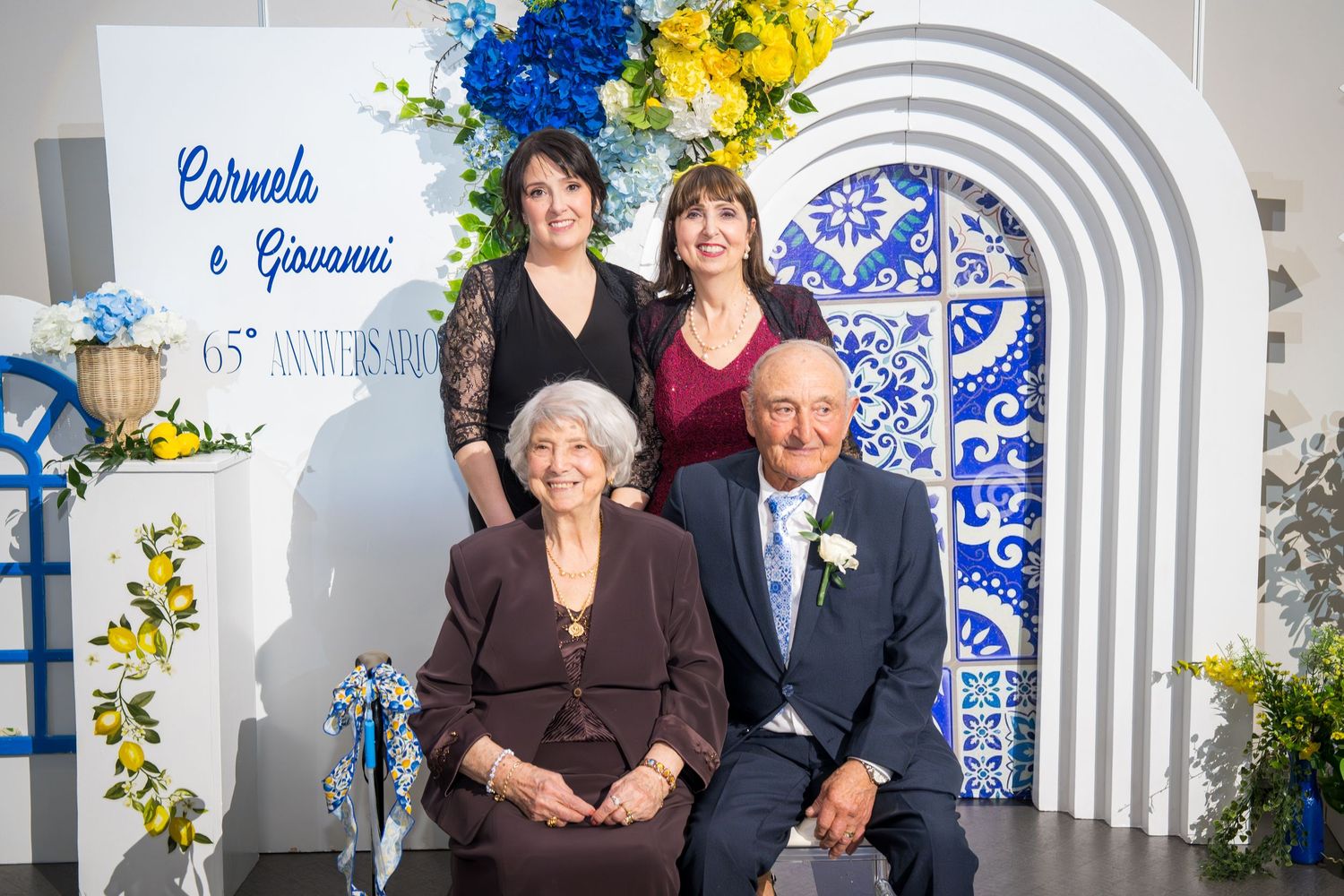 Carmela and Giovanni posing with two women at their 65th wedding anniversary celebration with floral arch backdrop.