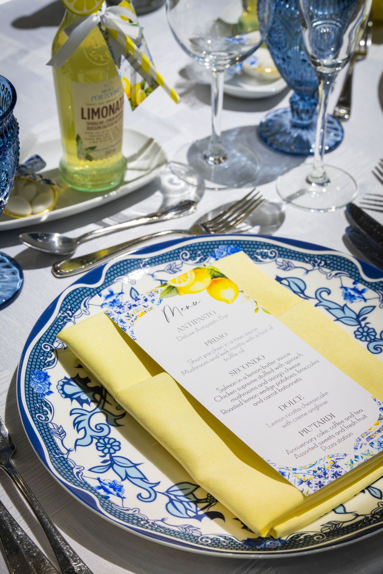 Blue and white floral china place setting with yellow linen napkin, lemon-themed menu card, and Limonata bottle on white tablecloth.
