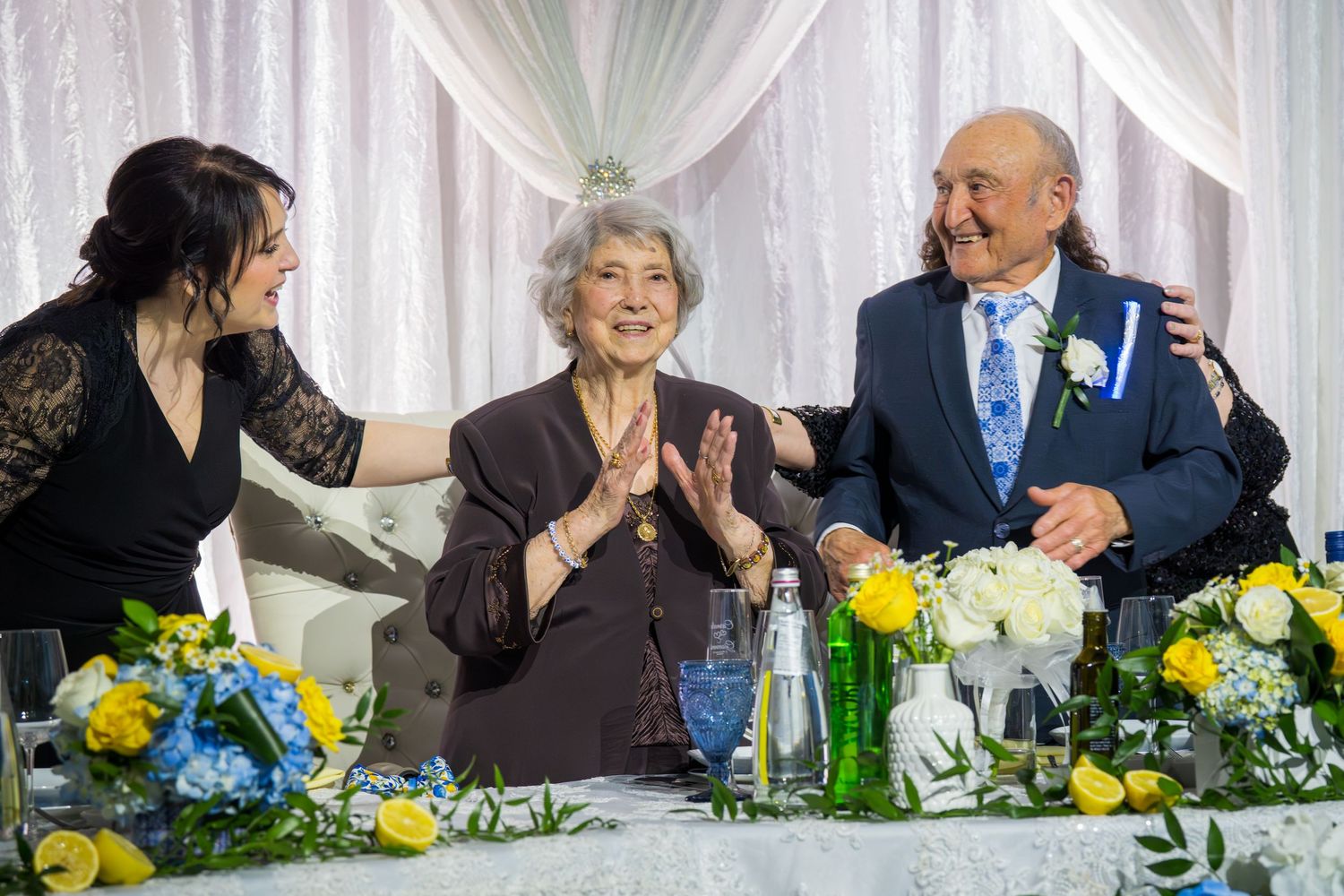 Joyful elderly woman clapping at anniversary dinner table surrounded by family members with yellow and blue floral decor.
