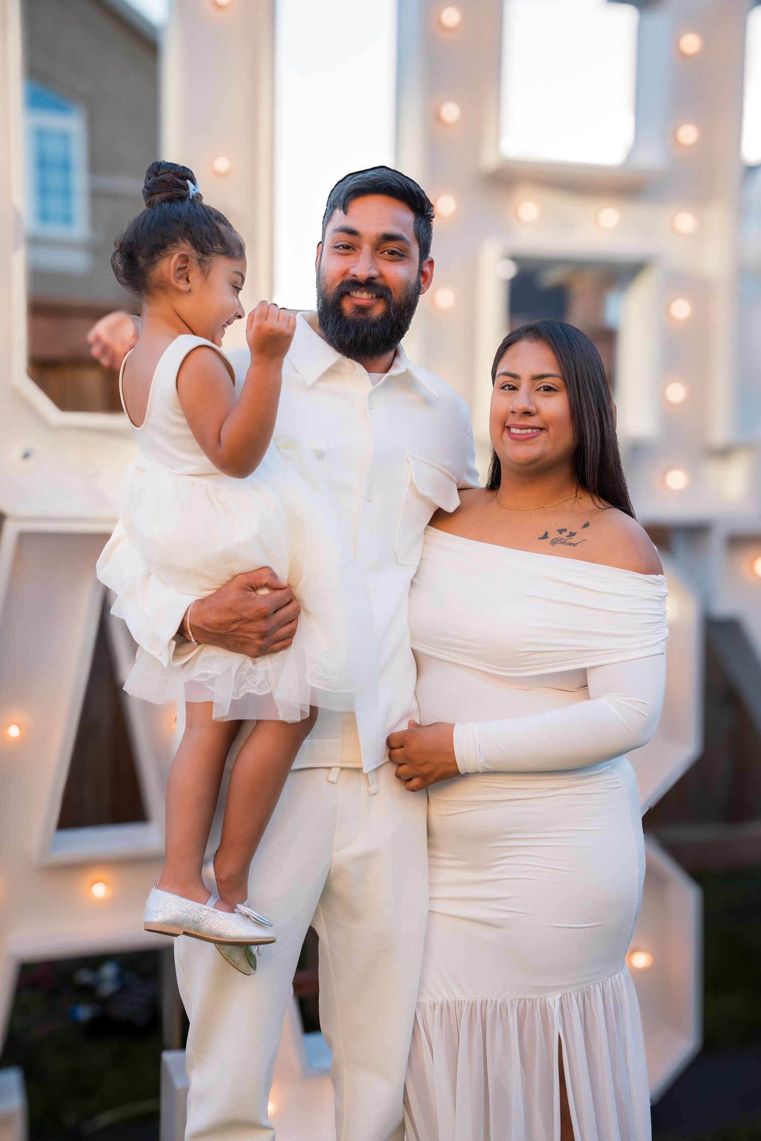 A bearded man in white holds a toddler girl while standing beside a pregnant woman at an outdoor gender reveal party.