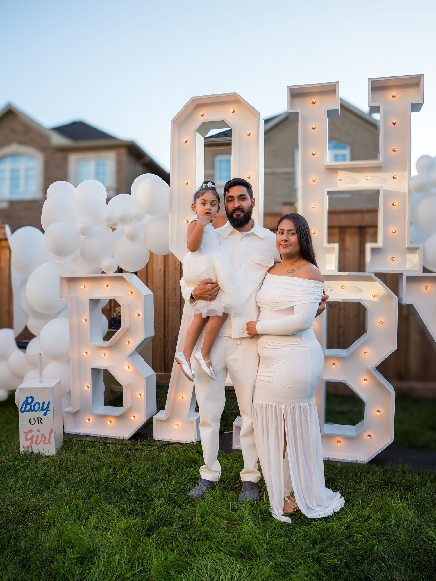 A family in white outfits poses before an illuminated OH BABY sign at a gender reveal party.