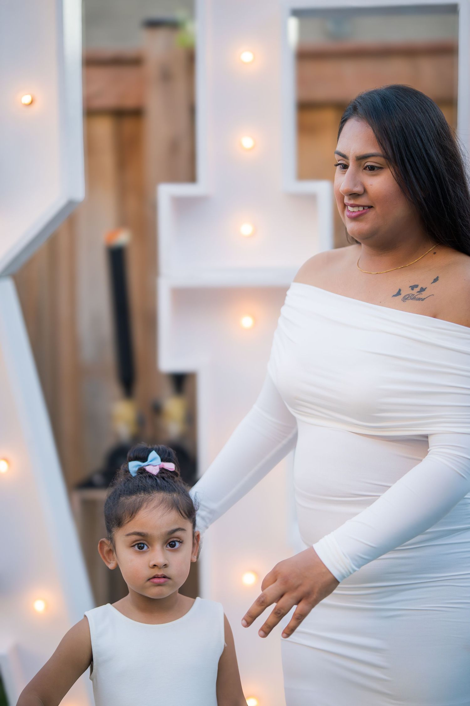 Smiling pregnant woman in white dress standing with young girl in front of lit decorative display.