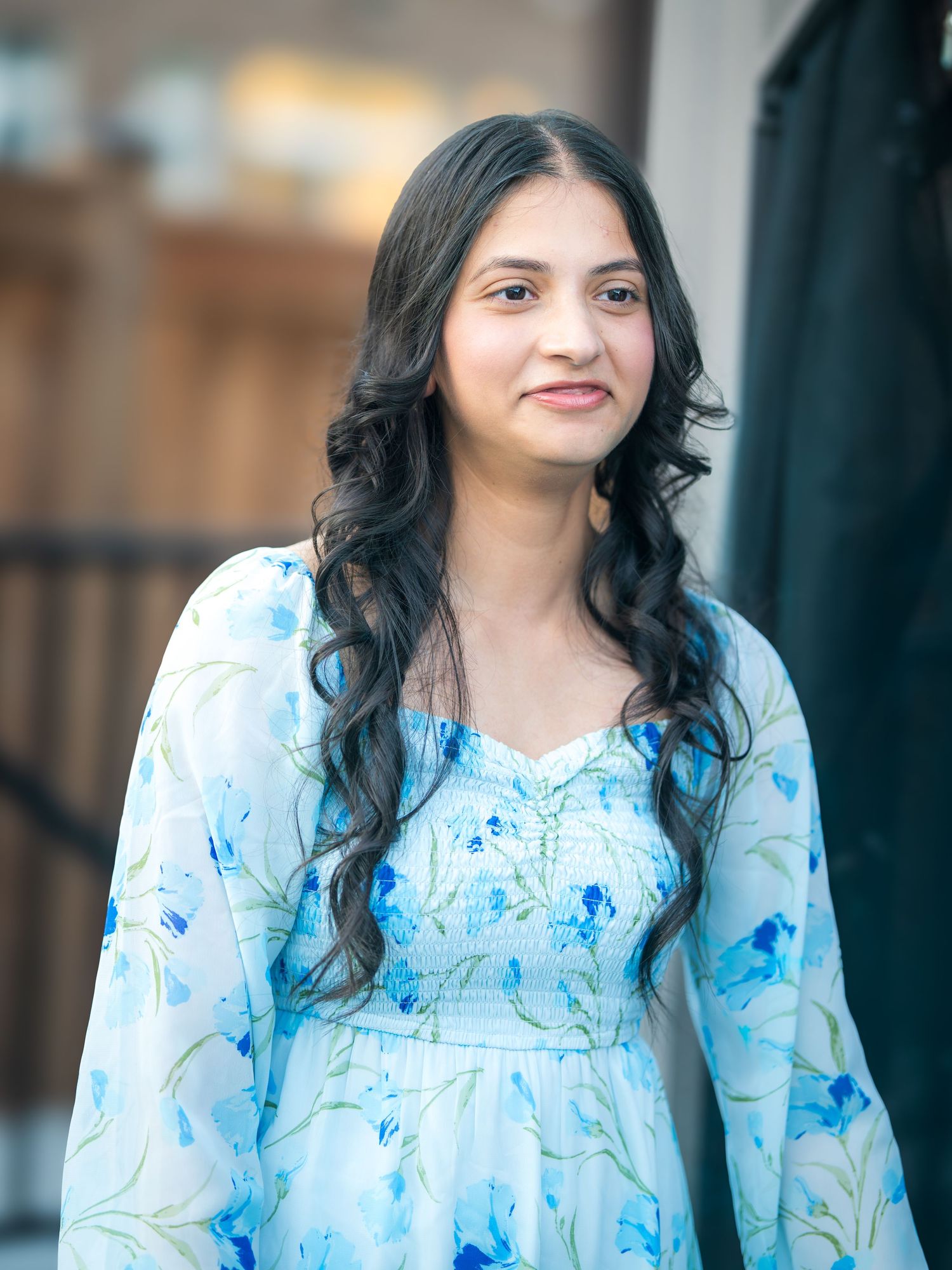 A young woman with long dark hair smiles in a floral blue dress at an outdoor event.