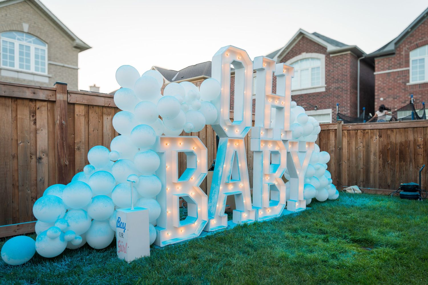 Illuminated OH BABY marquee letters surrounded by white and blue balloons at dusk in a backyard.