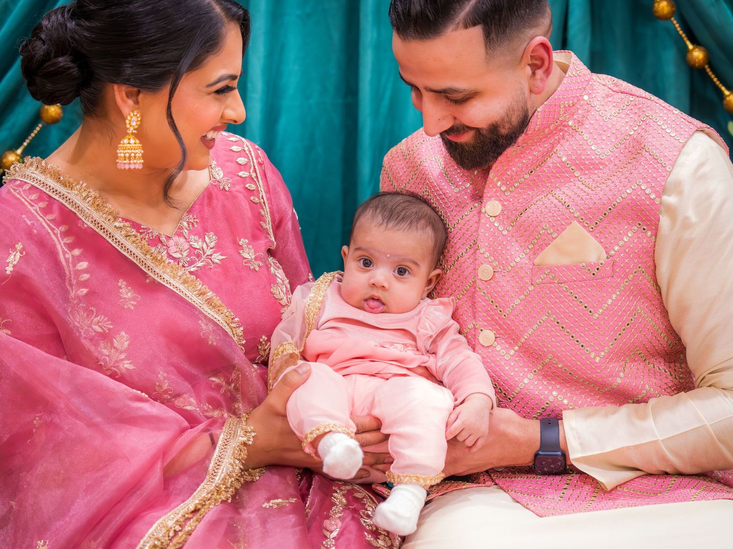South Asian parents in pink ethnic wear lovingly gaze at newborn baby at floral celebration event.