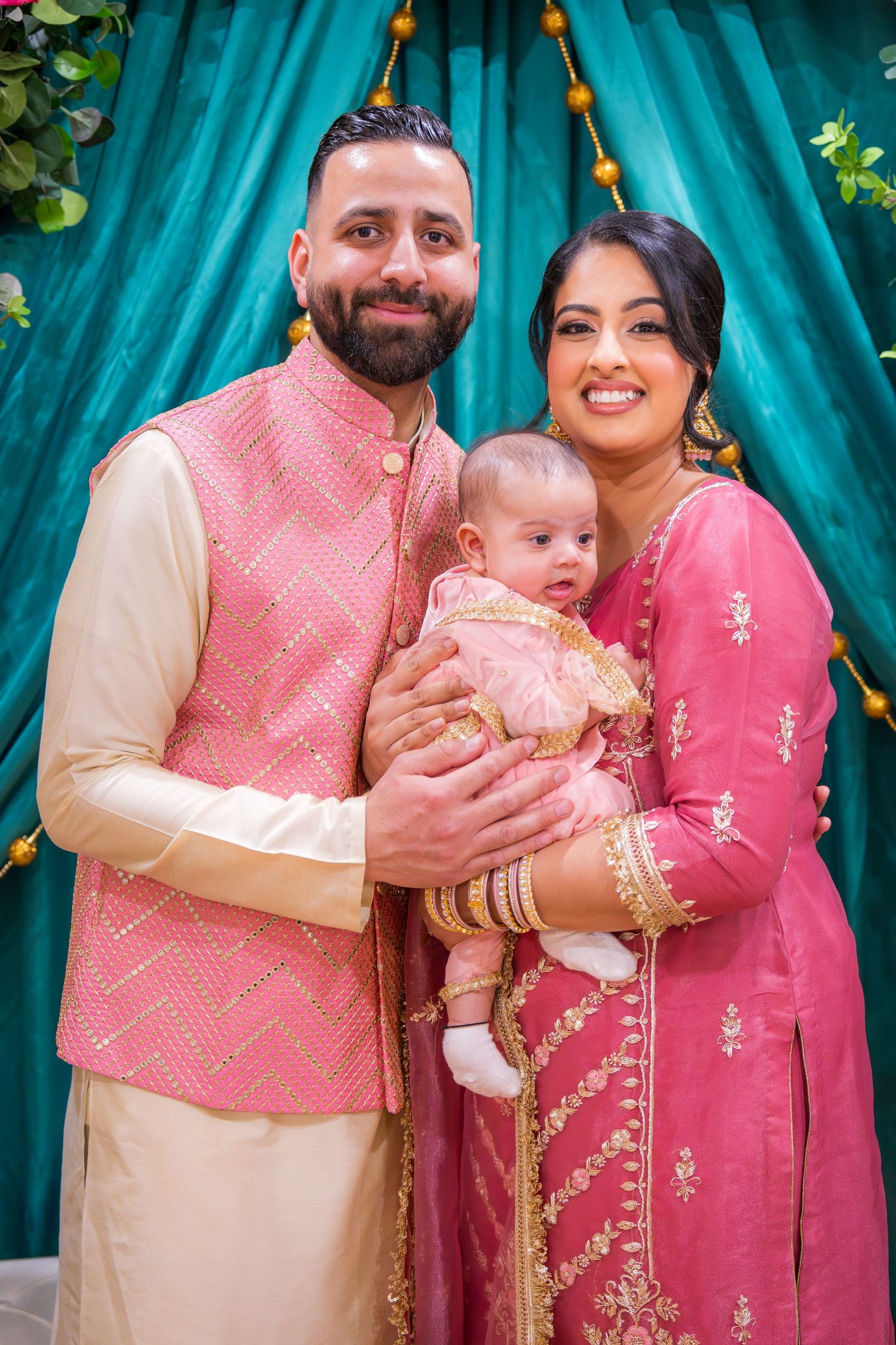 Couple in matching pink Indian outfits holding their baby at a festive South Asian celebration.