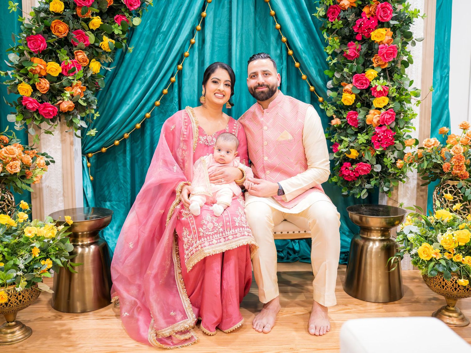 Indian family in pink traditional clothing seated with newborn baby before teal drape and vibrant floral arrangement.