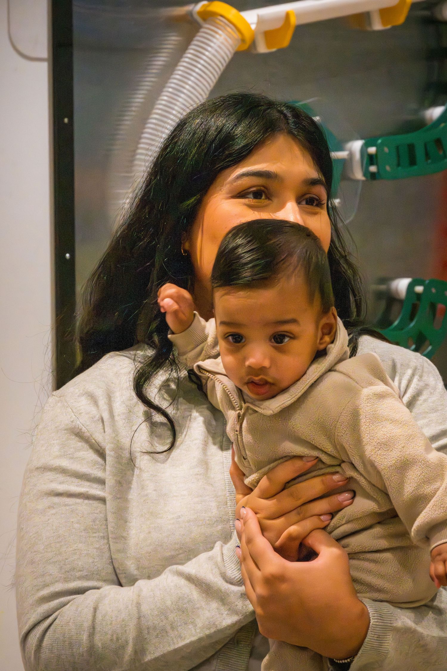 Young woman with long dark hair holding a baby in a grey sweater in an industrial setting.