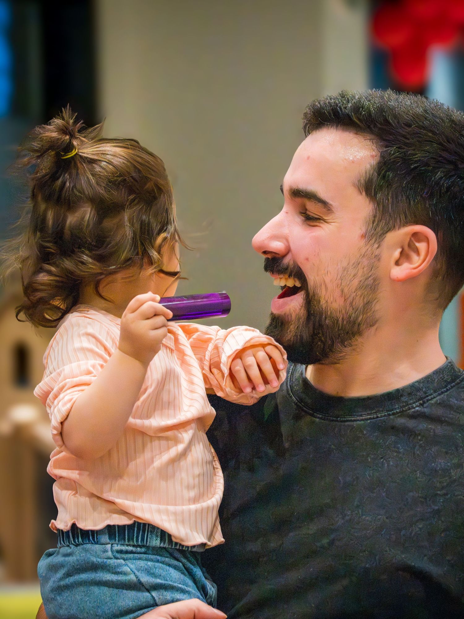 A bearded man laughs as a baby girl playfully holds a purple toy near his mouth.