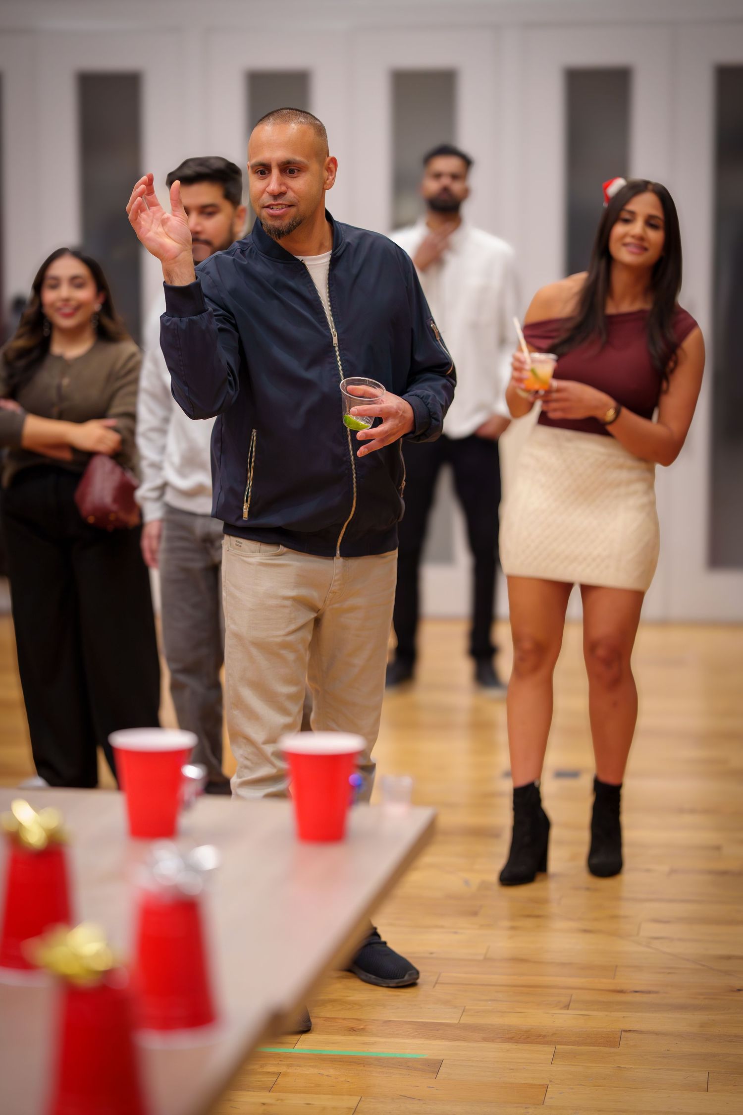 Party guests play a drinking game with red plastic cups arranged on a wooden floor.