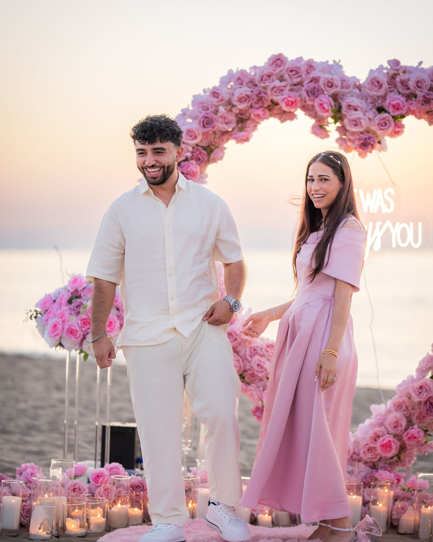 A beach engagement scene with pink floral decorations and couple in coordinated white and pink outfits.