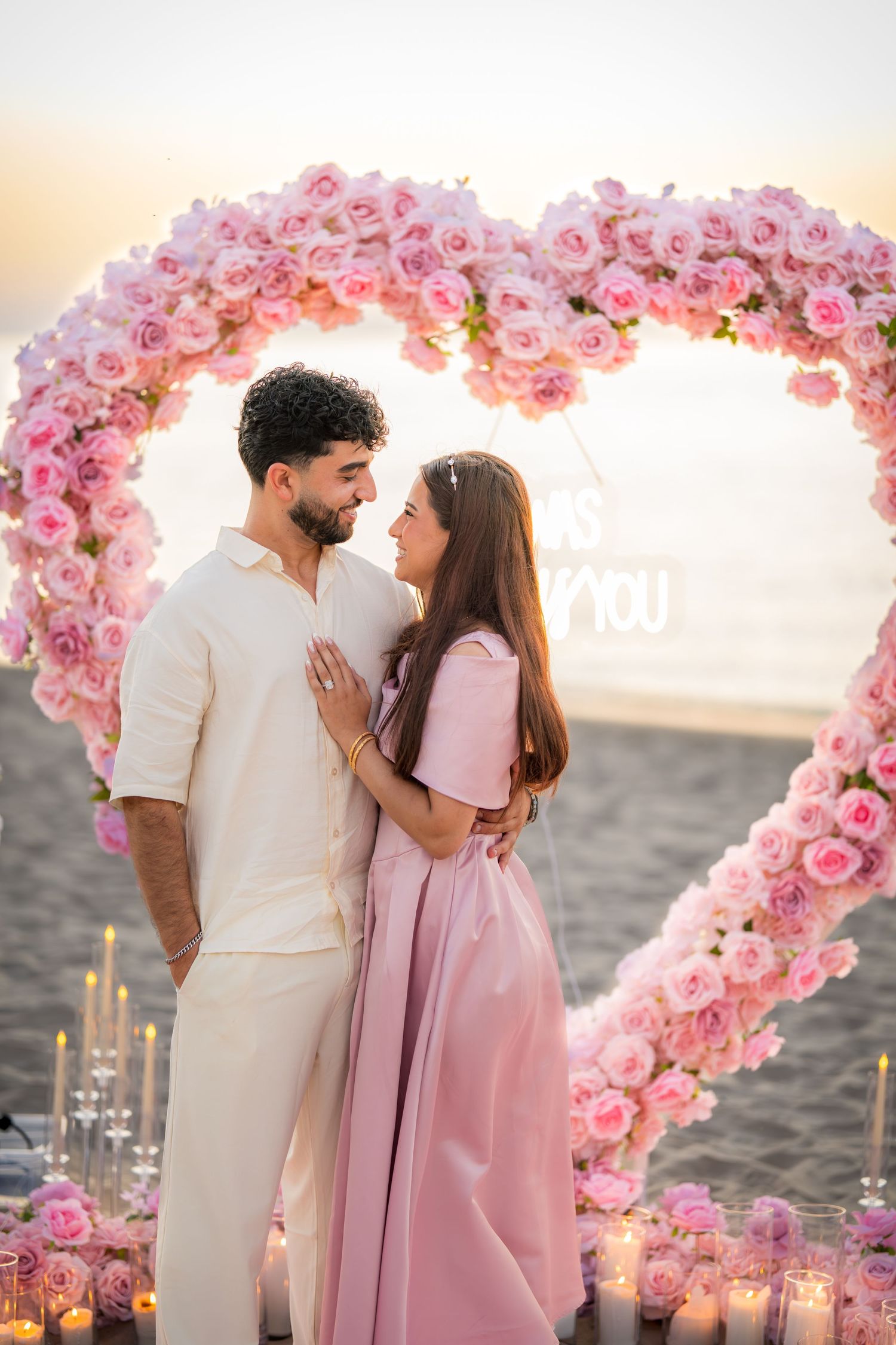 Romantic beach proposal scene with pink floral heart arch at sunset.