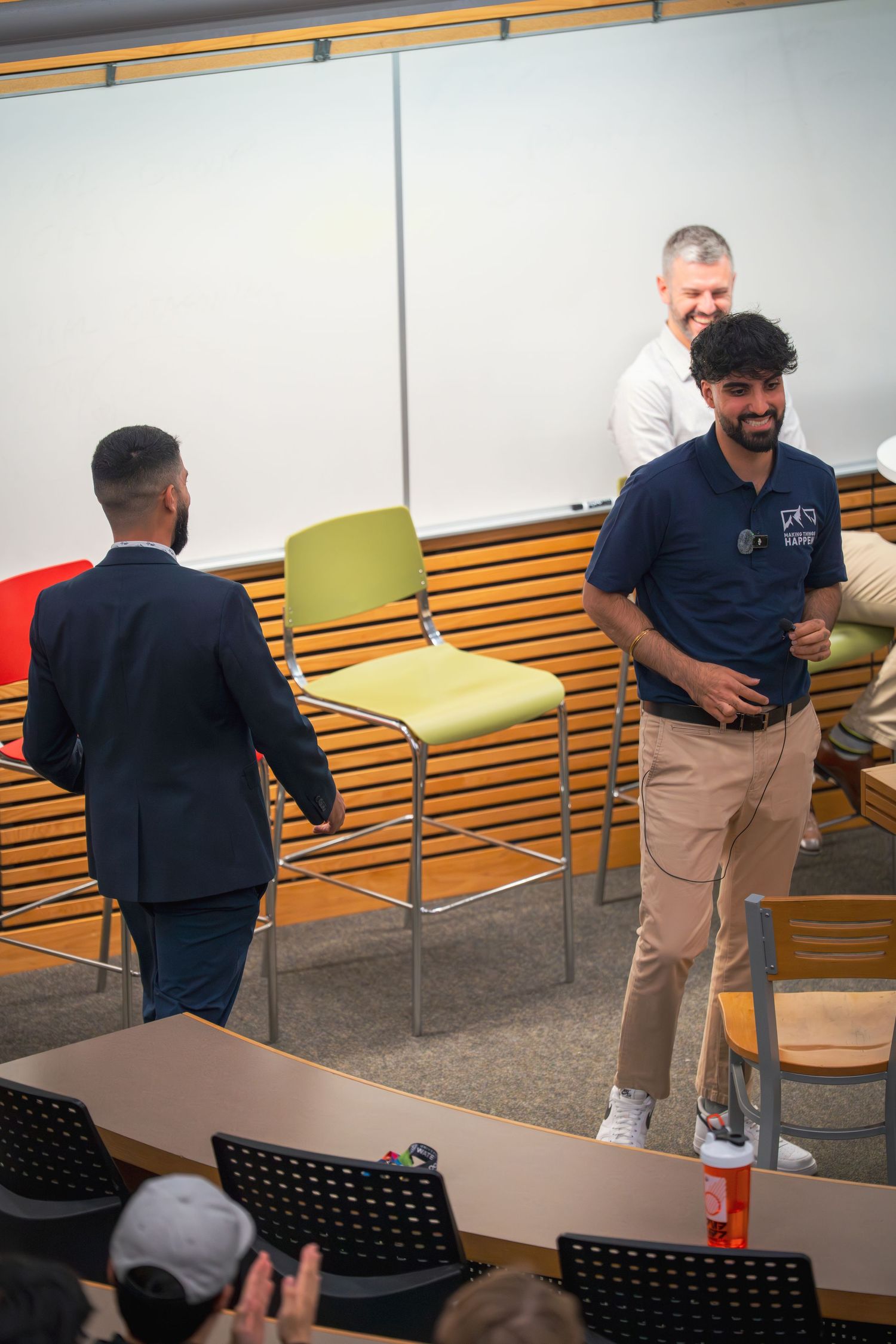 A presenter in a navy polo shirt and khaki pants gestures while speaking in a classroom setting.