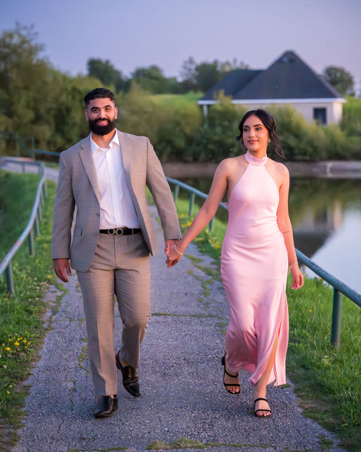 Two people stroll together on a gravel path beside a lake, wearing formal attire in pink and tan.