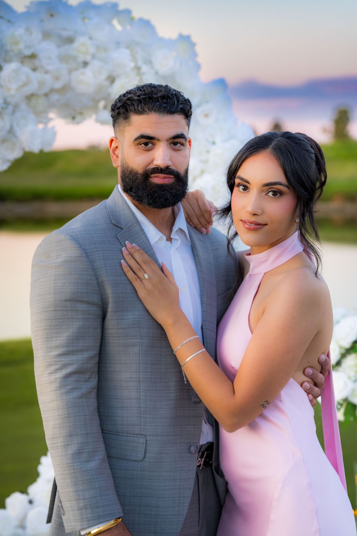 A couple in pink and grey formal attire poses together under a floral arch at sunset.