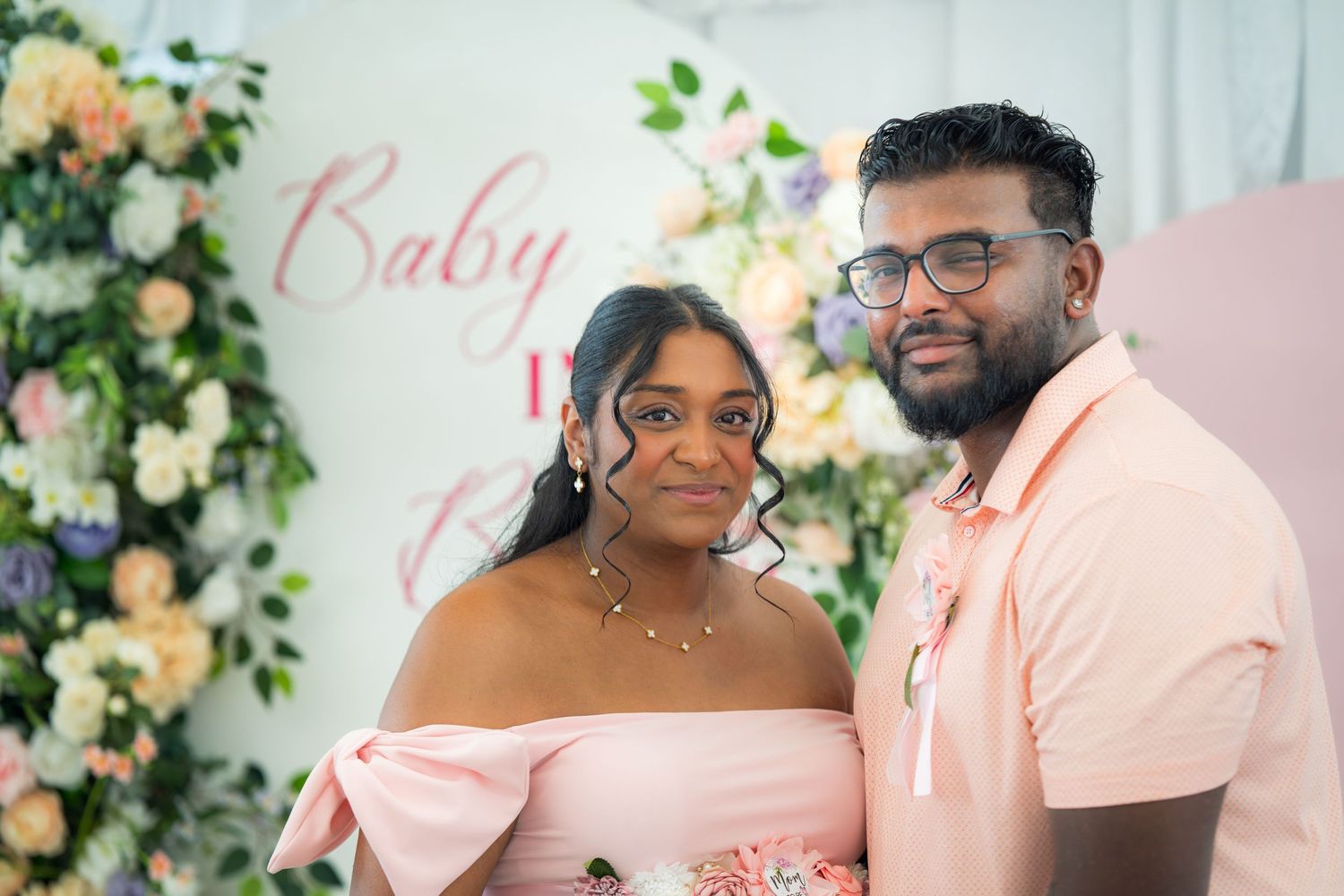 Baby shower photo featuring couple in matching pink and white outfits against floral backdrop.
