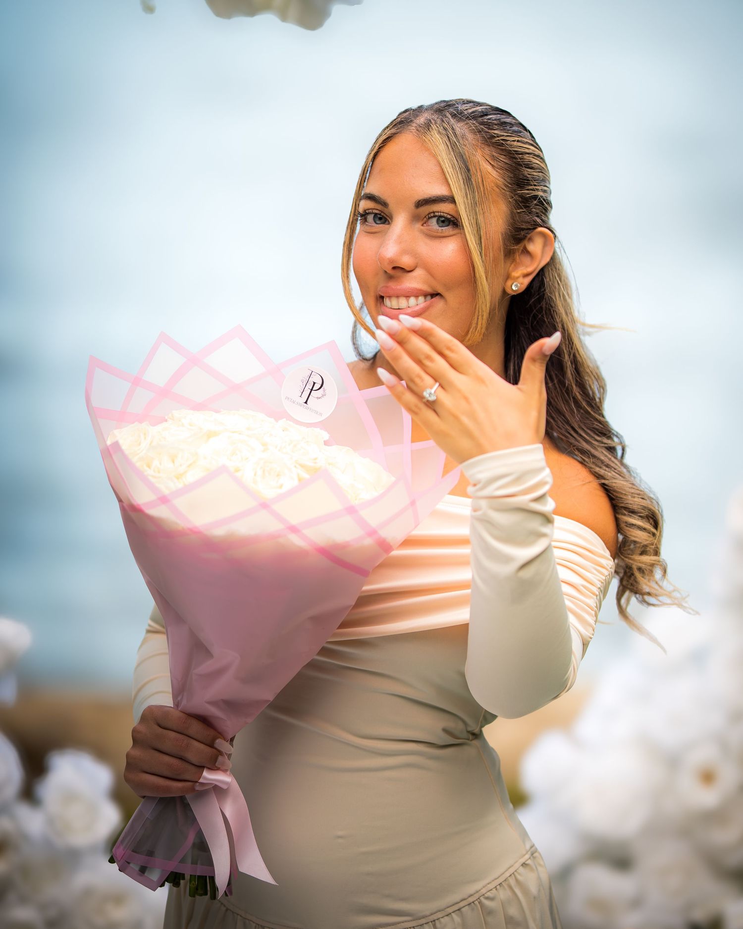 Person holding a pink-wrapped bouquet shows off a ring while posing outdoors.
