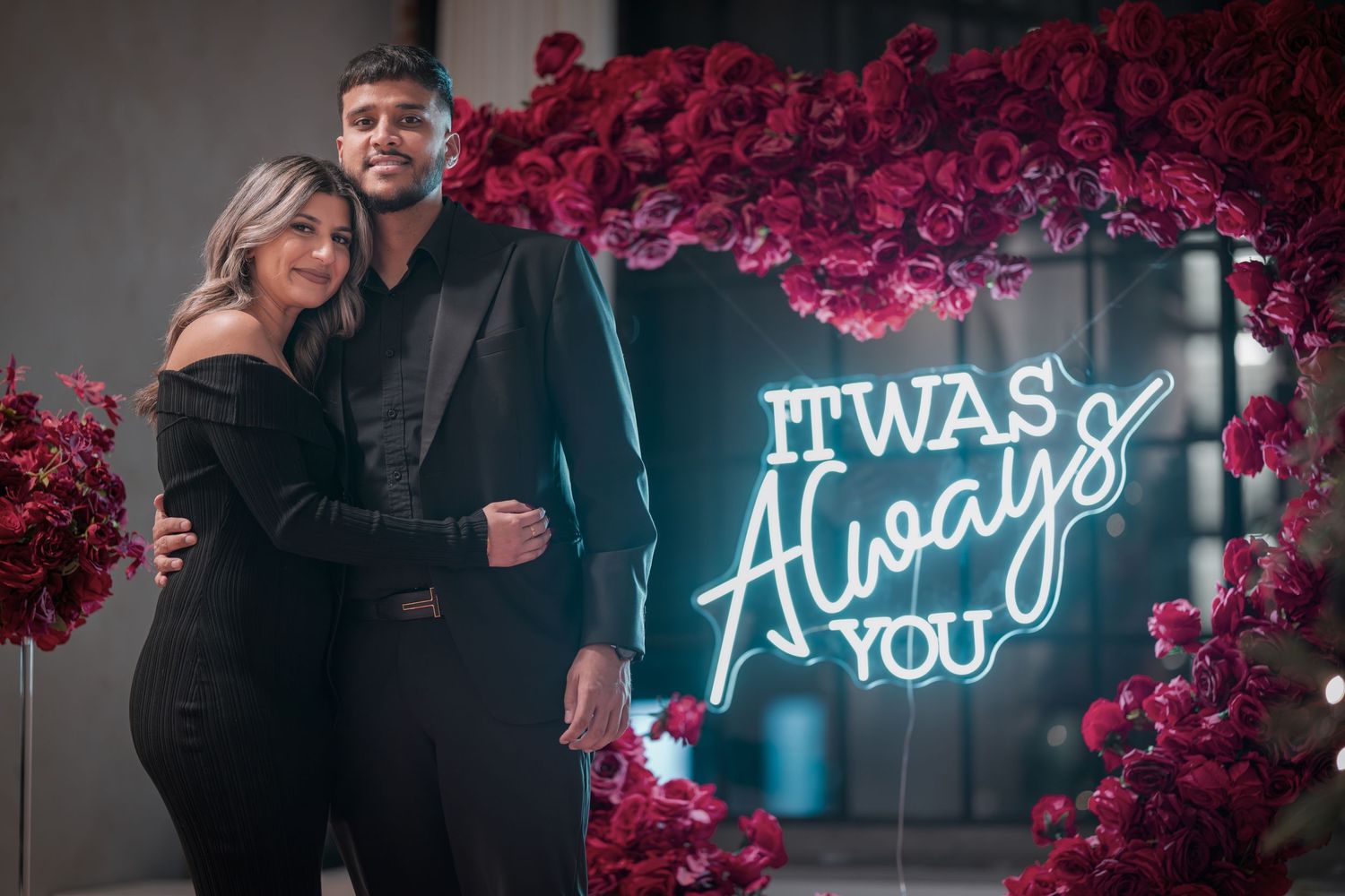 Neon sign reading 'It was you' glows against wall of red roses.