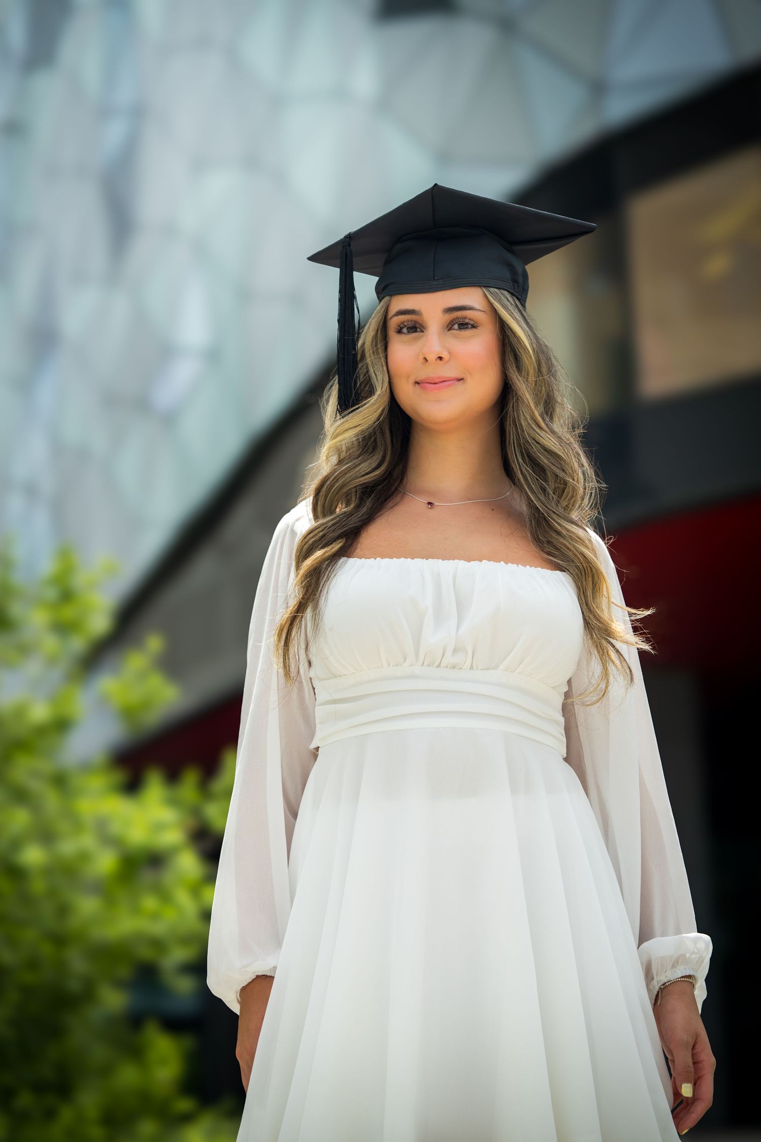 Graduate in flowing white dress poses in front of modern architecture.