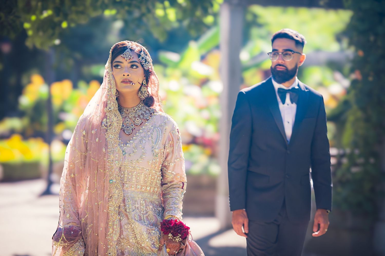 Newlyweds in traditional South Asian and Western fusion attire photographed in sunny outdoor garden.