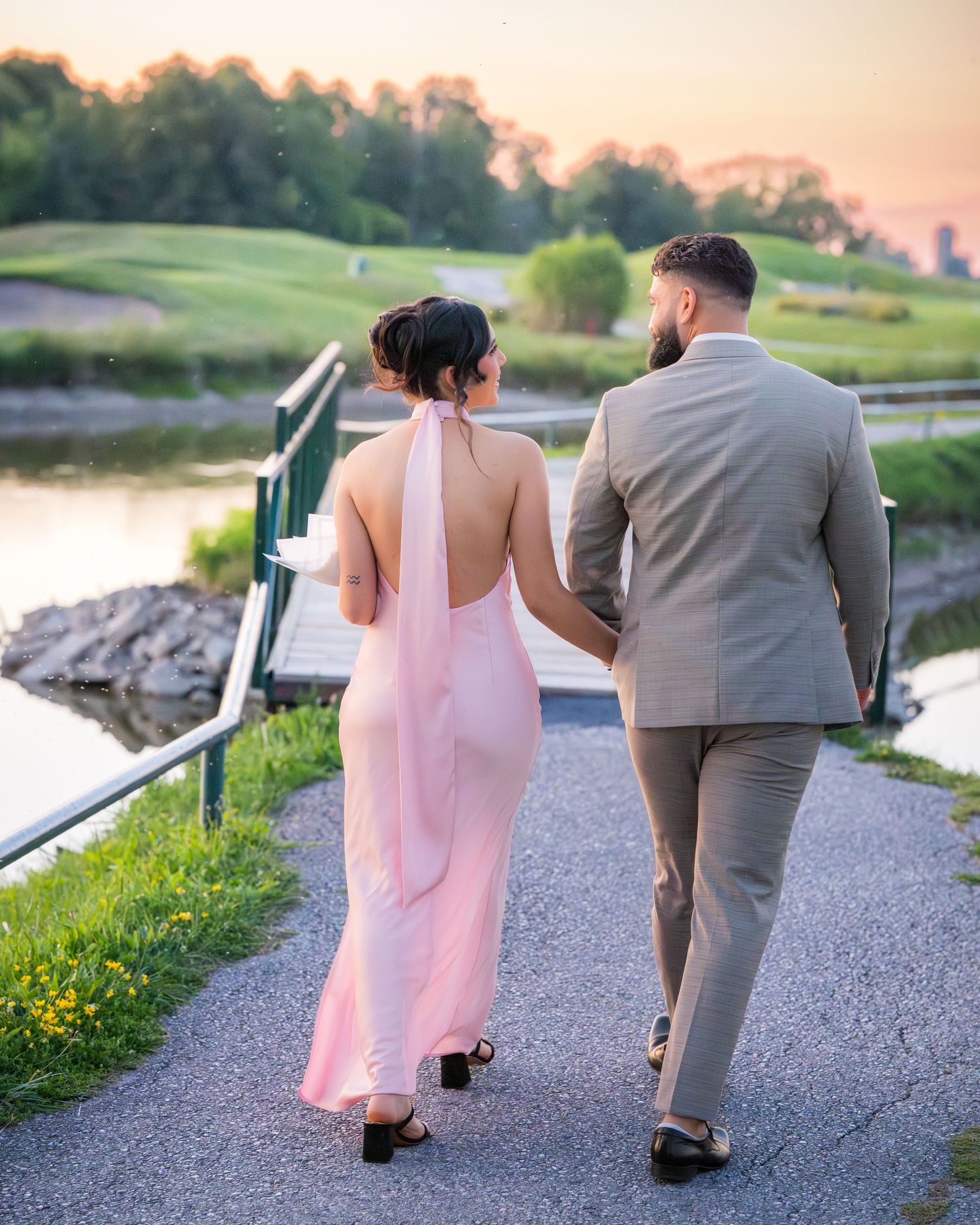 A couple in pink dress and grey suit walks along a waterside path at sunset.