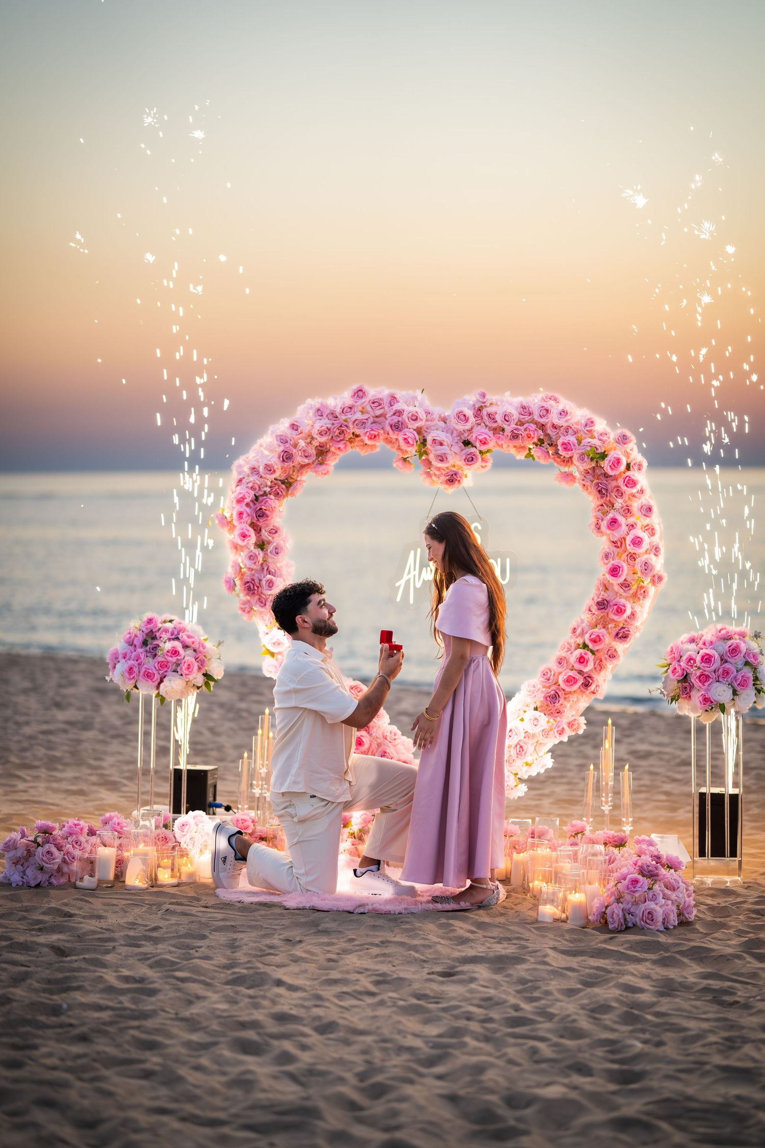 Romantic beach proposal with sparkling lights and pink floral heart arch during sunset.