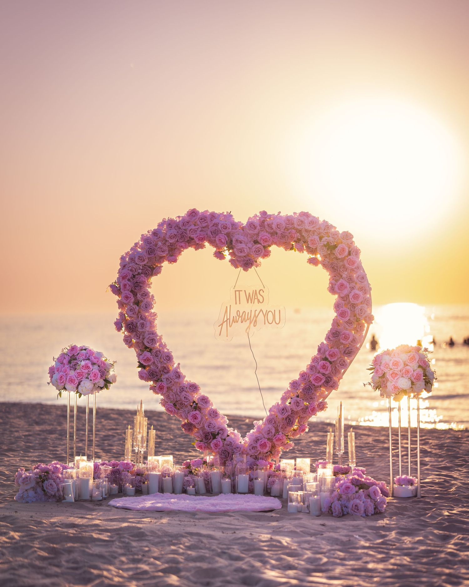 Heart-shaped floral arch with pink and purple flowers creates a romantic beach setting at sunset.