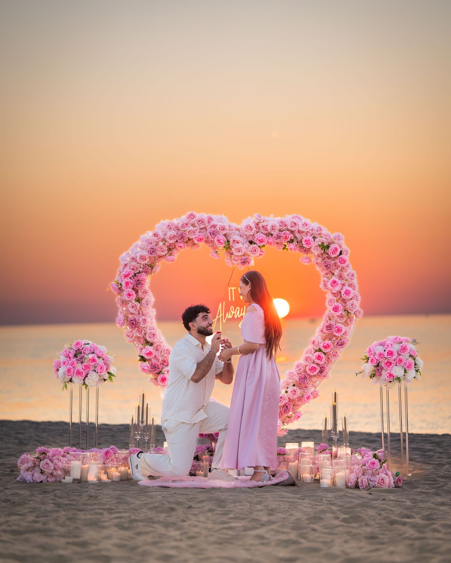 Romantic beach proposal setup with pink floral heart arch and candles at sunset creating a dreamy atmosphere.