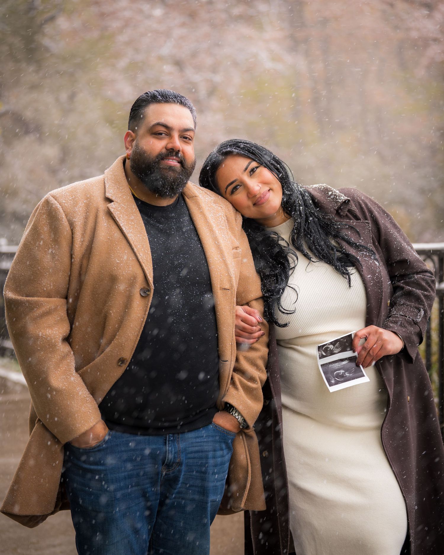 Couple embracing outdoors during winter snowfall while holding ultrasound photo.