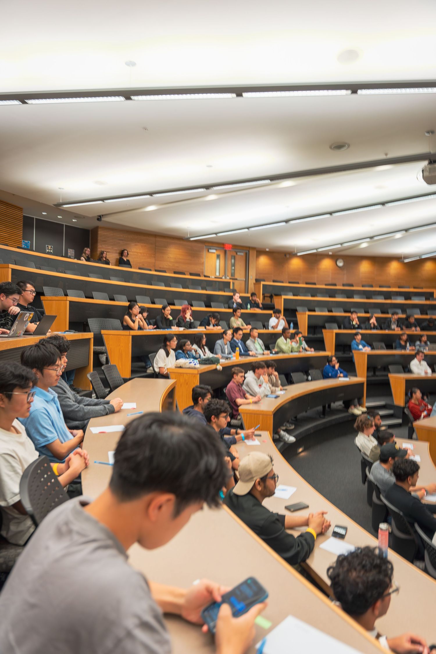 Students sit attentively in tiered lecture hall seating during a presentation or class.