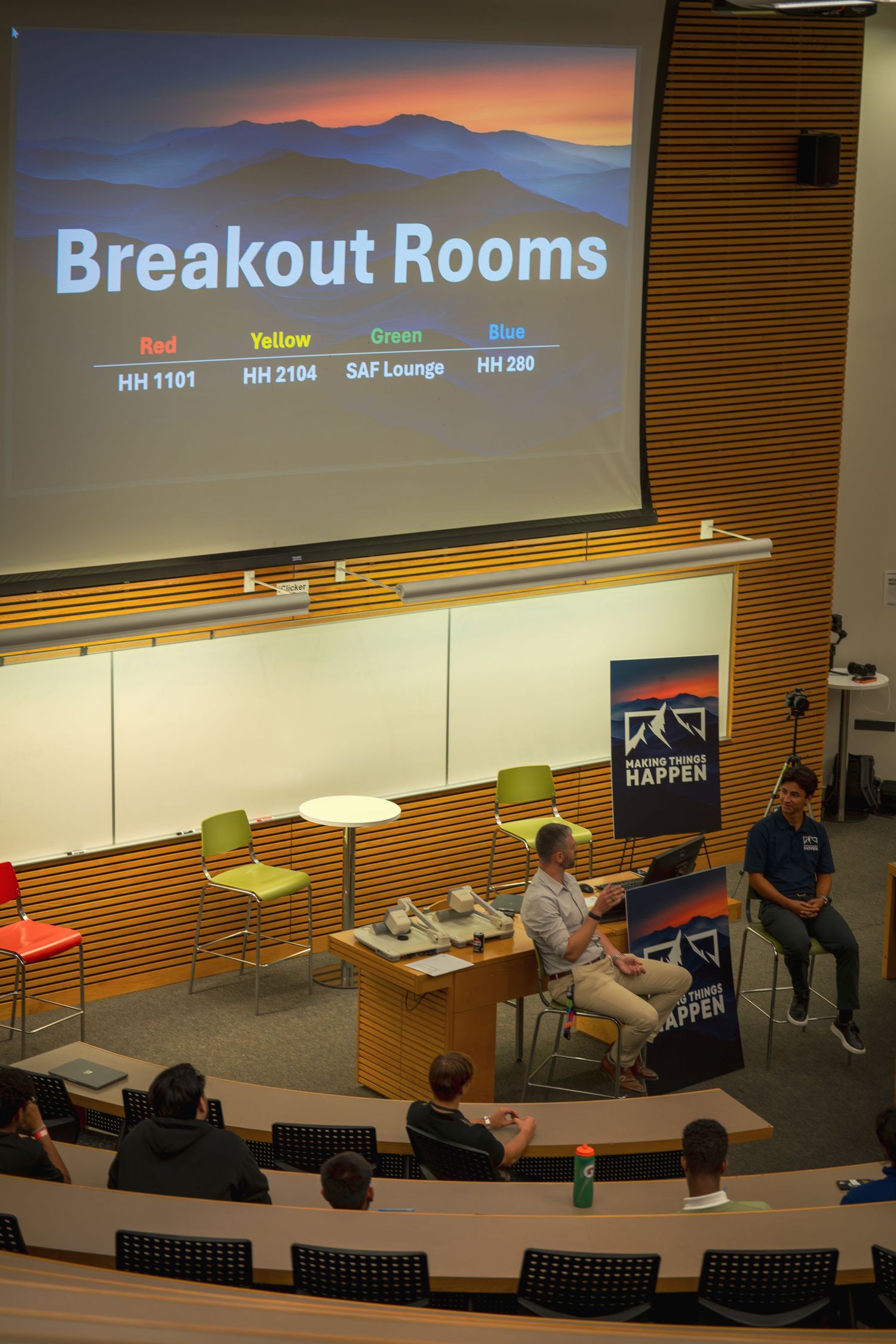 Breakout rooms presentation displayed on screen in tiered lecture hall with audience.