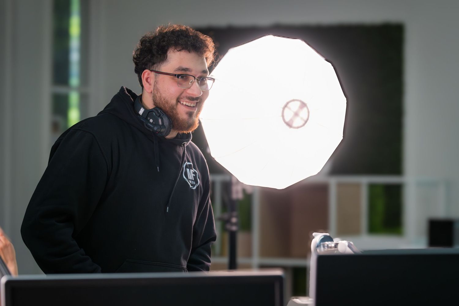 Young man with headphones and glasses smiling behind DJ equipment with a large softbox light in modern office.